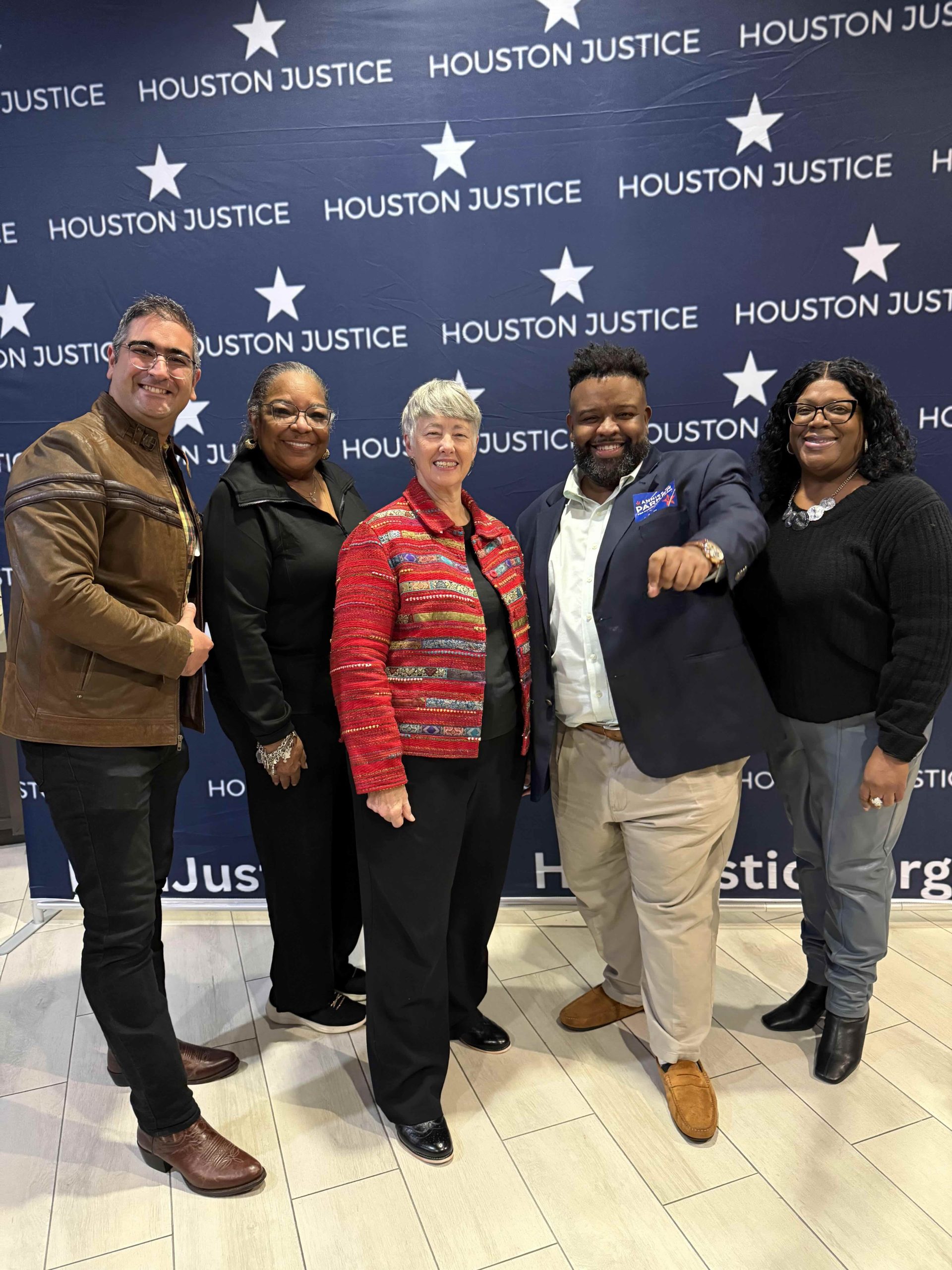 Un grupo de cinco personas posando frente a un fondo que dice "HOUSTON JUSTICE", con sonrisas y atuendos variados en un ambiente de celebración.