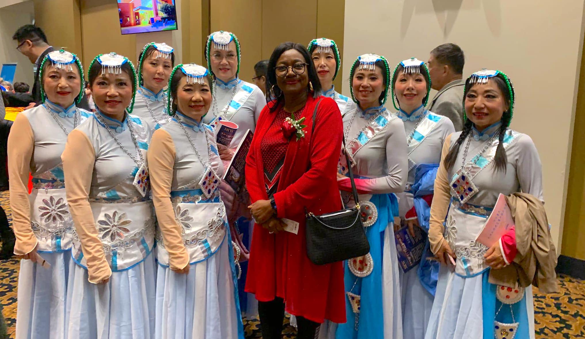 Celebración Cultural con Grupo de Baile:::Un grupo de mujeres vestidas con trajes tradicionales posan para una foto, acompañadas por una mujer en un vestido rojo, en un evento cultural.