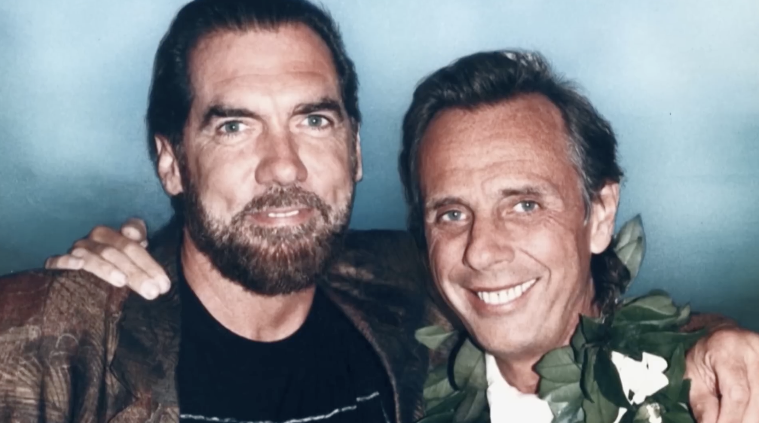 Dos hombres sonriendo y posando juntos, uno con una corona de flores y el otro con una chaqueta colorida, fondo azul.