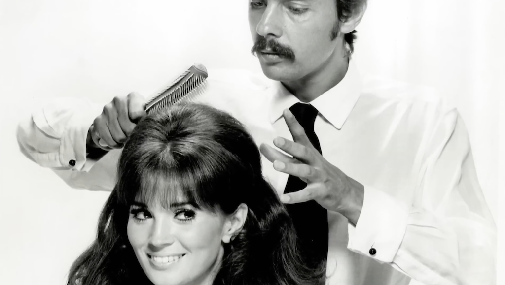 Un hombre con bigote peina a una mujer con cabello largo y voluminoso, sonriendo a la cámara.