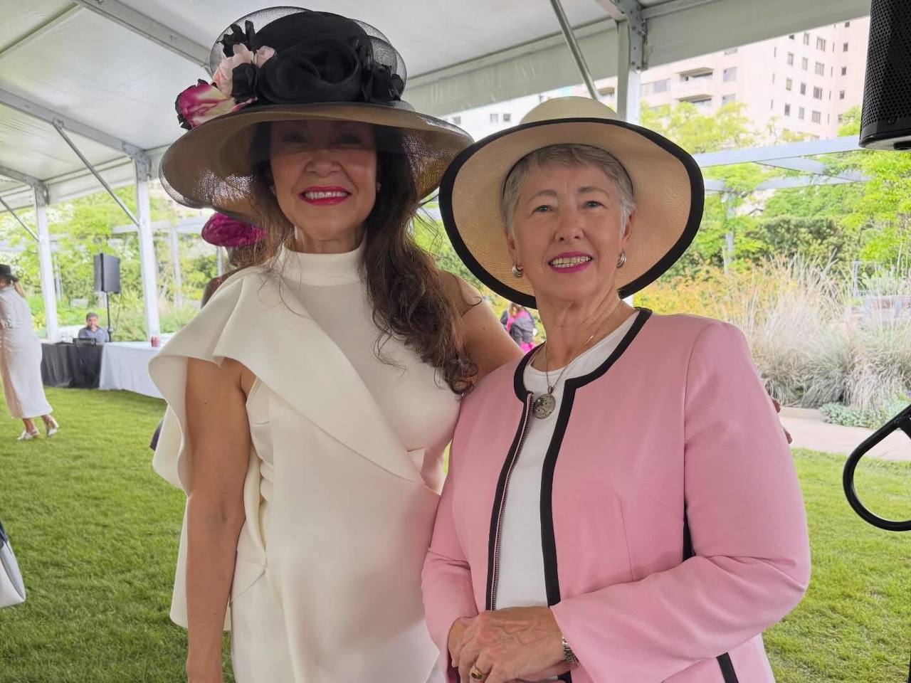 Annise Parker posando con una elegante asistente en vestido blanco y sombrero negro en The Hats in the Park Gala
