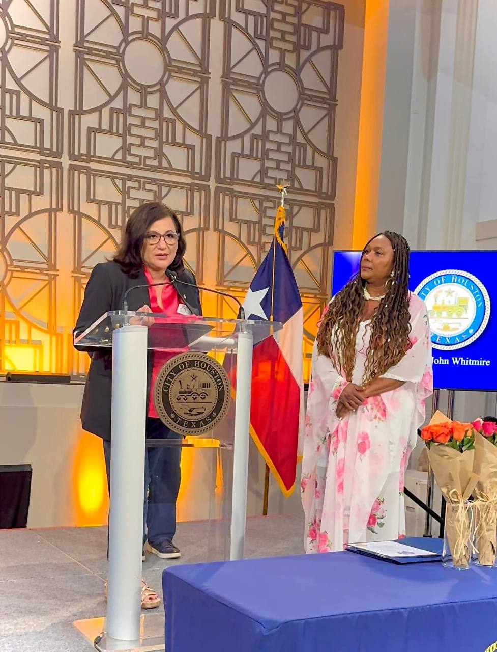 Daysi Marin hablando en el podio del City of Houston con la Dra. Sonia White a su lado y bandera de Texas de fondo