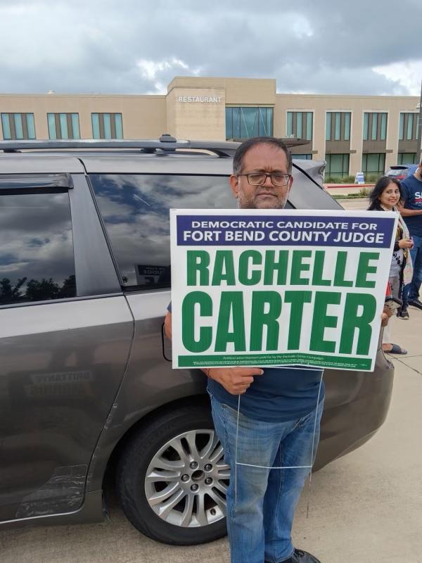 Hombre sosteniendo letrero de Rachelle Carter candidata democratica para Fort Bend County Judge junto a una van